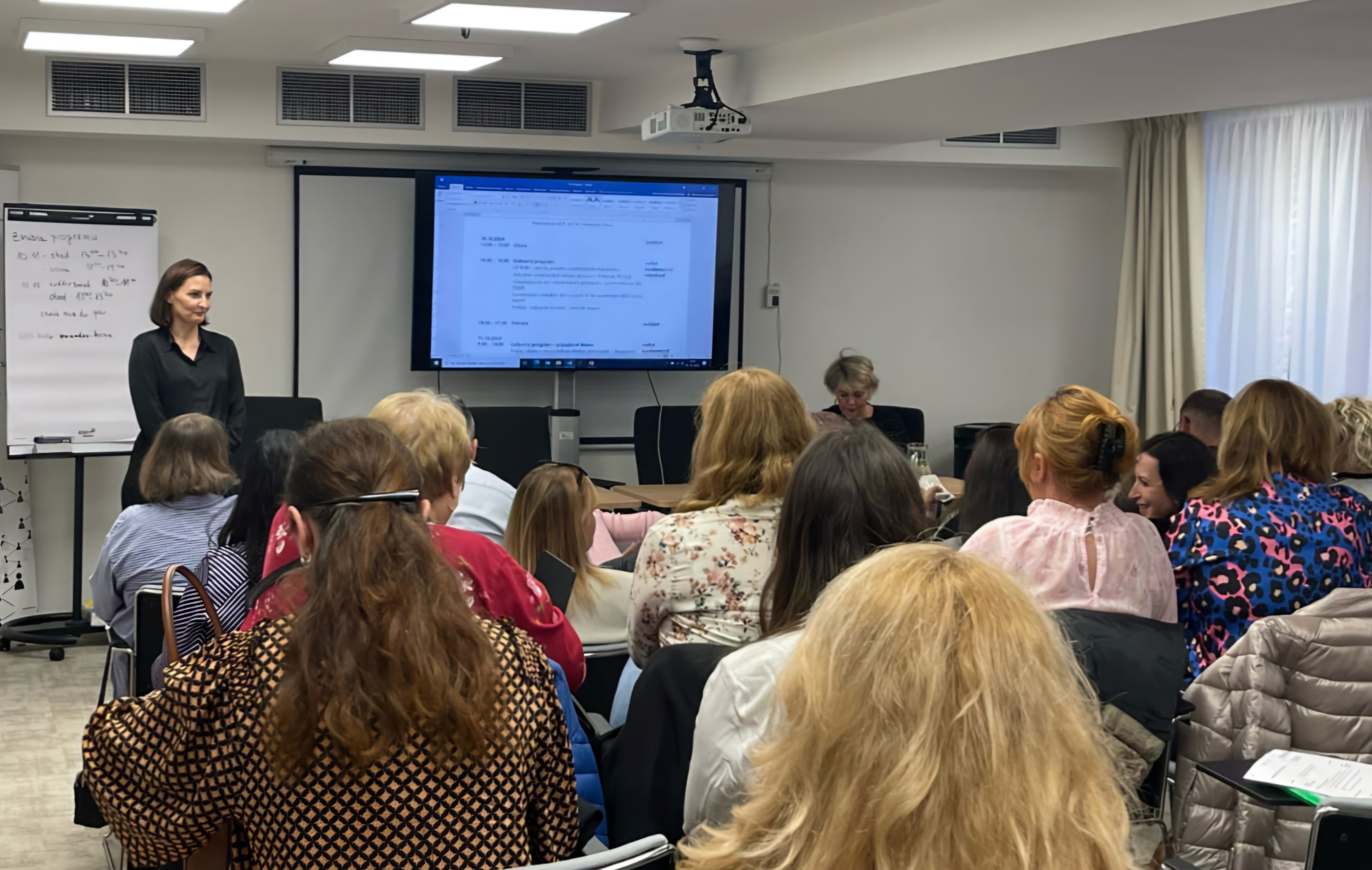 foto 04 | WebJET CMS Účastníci porady sediaci na stoličkách, pred nimi vedľa obrazovky s prezentáciou stojí prednášajúca riaditeľka Nadežda Vrtielová Prečinská